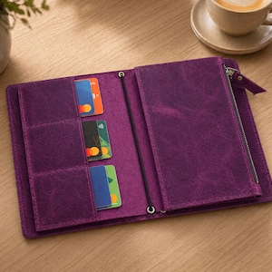 May include: Open purple leather wallet with multiple card slots and a zipped compartment. The wallet is open, revealing several credit cards in various colours. A black elastic band and metal accents secure the wallet. A cup of coffee sits in the background.