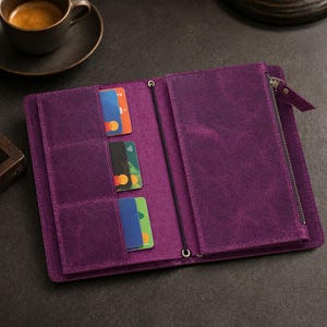 Può includere: Portafoglio in pelle viola aperto con più fessure per carte e uno scomparto con cerniera. Il portafoglio è aperto, rivelando diverse carte di credito di vari colori. Un elastico nero assicura il portafoglio. Una tazza di caffè è sullo sfondo.