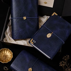 May include: Navy blue leather journals with gold accents, including a flower and leaf charm. The journals are closed with elastic bands and are presented in a black box with a decorative card.