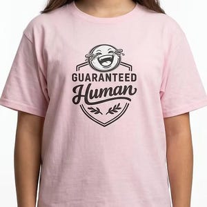 May include: A light pink t-shirt featuring a black and white graphic. The graphic includes a laughing emoji, the words "GUARANTEED" above "Human", and a shield design with leaves.