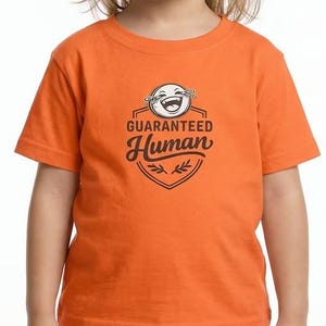 May include: Orange t-shirt with a graphic that reads "GUARANTEED Human". The graphic features a laughing emoji and a shield design. The shirt is short-sleeved and appears to be made of cotton.