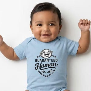 May include: A light blue baby onesie with the text "GUARANTEED Human" in a black and white graphic. The onesie features a smiling face graphic. The baby is smiling and has their arms raised.