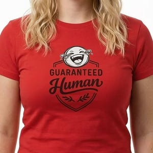 May include: A red t-shirt featuring a graphic with a laughing emoji and the words "GUARANTEED Human" in a shield design. The shirt is short-sleeved and made of a soft material.