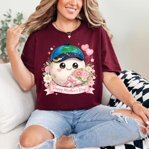 May include: Burgundy t-shirt featuring a cartoon character with a baseball cap and floral accents. The character has a smiling face and is surrounded by flowers and a banner that reads "Happy Mother's Day!"