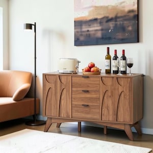 May include: A wooden sideboard featuring three central drawers with dark handles, flanked by two cabinets with arched door panels. On top sits a cream-colored toaster, a woven basket containing red apples, and three wine bottles with two glasses.