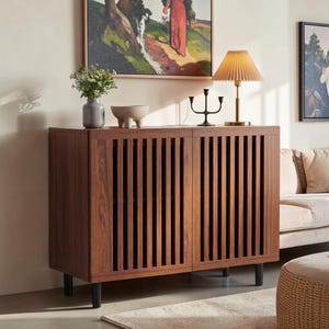 May include: A rectangular wooden cabinet with two doors featuring vertical slats, supported by four black legs. On top sits a gray vase with greenery, a small beige bowl, a black metal candle holder, and a lamp with a pleated beige shade.