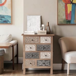 May include: A wooden chest of drawers featuring seven drawers with varied embossed floral patterns in shades of white, gray, and natural wood. Each drawer is equipped with a small, dark, round knob. The top surface holds a white rectangular frame.