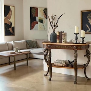 May include: A dark wood console table with ornate carved legs featuring animal motifs, a lower shelf holding two books and a decorative object, and a top surface displaying a dark vase with dried branches, three books, and two white pillar candles.