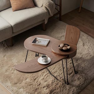 May include: A unique, wooden coffee table with a built-in chair. The table has a light brown finish, with a small round tray and a lower, curved surface. A cup of coffee and a book are on the table.