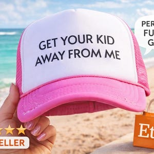 May include: A white and pink trucker hat with the text "GET YOUR KID AWAY FROM ME" in black. The hat has a pink brim and mesh back. A speech bubble says "PERFECT FUNNY GIFT!" An orange Etsy bag is in the background.
