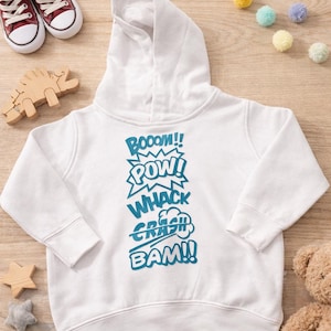 May include: White hooded sweatshirt with blue comic book sound effects: BOOOM!! POW! WHACK CRASH BAM!! Red canvas sneakers and wooden toys are arranged around the sweatshirt on a wood grain surface.