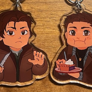 May include: Two acrylic keychains featuring cartoon illustrations of characters. One keychain depicts a character holding a knife, the other shows a character eating pie. Both characters are wearing brown jackets.
