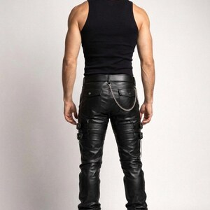 May include: Black leather pants with multiple buckles and a silver chain detail. The pants have a ribbed design on the knees and pockets on the back. Paired with a black tank top and black boots.