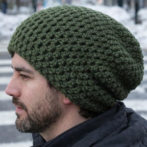 May include: Olive green crochet beanie hat. The hat is made of thick yarn with a textured stitch pattern. The beanie is a slouchy style, with a relaxed fit. The hat is shown on a person's head, with a blurred background.