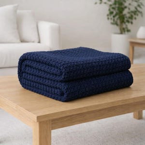 May include: A folded navy blue blanket with a textured weave sits on a light wood table. The blanket is made of a soft material and is folded neatly. The background includes a white sofa and a potted plant.