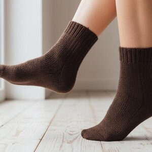 Pu&ograve; includere: Un paio di calzini marroni a maglia a coste. I calzini sono indossati sui piedi di una persona, con un piede leggermente sollevato da un pavimento in legno chiaro. I calzini hanno un aspetto spesso e strutturato.