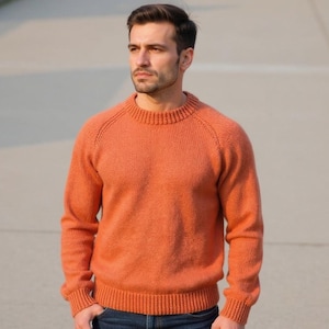 Puede incluir: Un suéter naranja de cuello redondo. El suéter tiene cuello, puños y dobladillo acanalados. El suéter de manga larga está hecho de un material suave y texturizado. El modelo lleva vaqueros azules.
