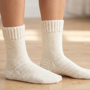 Peut inclure: Une paire de chaussettes en crochet blanc cassé. Les chaussettes ont un bord-côte et un motif ajouré texturé. Les chaussettes sont présentées sur une surface en bois.