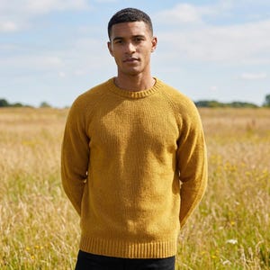Könnte beinhalten: Senfgelber Pullover mit Rundhalsausschnitt. Der Pullover hat lange Ärmel und gerippte Bündchen. Die Person steht in einem Feld mit hohem Gras unter blauem Himmel.