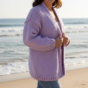 May include: A lilac-colored, chunky knit cardigan with long sleeves. The open-front sweater is made of a thick, textured yarn. The person is standing on a sandy beach with the ocean in the background.