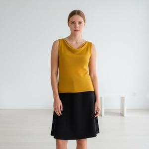 May include: A woman in a mustard yellow sleeveless top with a cowl neck and a black knee-length skirt. The outfit is simple and elegant, suitable for various occasions. The background is a minimalist white room.