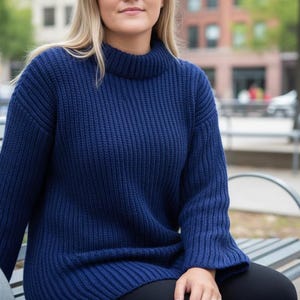 Può includere: Un maglione a collo alto in maglia grossa blu navy. Il maglione ha una trama a coste e una vestibilità ampia. Le maniche sono lunghe e lo scollo è un collo alto. La persona è seduta su una panchina di metallo.