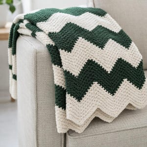 Könnte beinhalten: Eine handgehäkelte Decke mit einem Zickzackmuster in Dunkelgrün und Creme. Die Decke ist über den Arm eines hellgrauen Sofas drapiert und zeigt ihre Textur und ihr Design. Dieser gemütliche Überwurf ist ideal für die Inneneinrichtung.