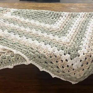 May include: A handmade crochet blanket in shades of white, beige, and green. The blanket has a scalloped edge and a textured pattern. The blanket is folded over a wooden surface.