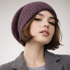 May include: A knitted, slouchy beanie in a deep purple hue. The hat has a textured stitch pattern and a relaxed fit. The model is wearing a grey blazer and a grey shirt.