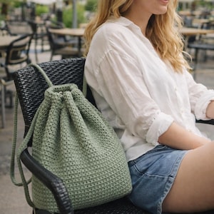 May include: A sage green crochet drawstring backpack with a textured stitch pattern. The bag has a rounded shape and thick, braided straps. The bag is sitting on a black chair.