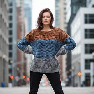 May include: A color-blocked sweater in brown, blue, and gray. The sweater has a round neckline and long sleeves. The model is wearing black pants. The background is a city street with tall buildings.