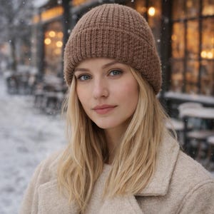 Puede incluir: Una mujer con un gorro de punto marrón y un abrigo beige. El gorro es de un color cálido y neutro, y el abrigo tiene un diseño clásico y entallado. El fondo muestra una escena nevada al aire libre con luces borrosas.