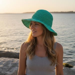 May include: A turquoise crochet sun hat with a wide brim. The hat is worn outdoors near the sea. The hat is a stylish accessory for sun protection. The hat is made of a textured yarn.