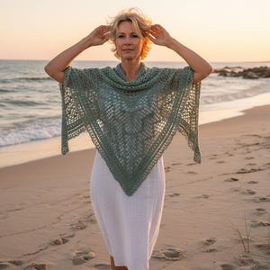 Peut inclure: Une femme porte un châle en crochet vert sauge et une robe blanche sur une plage au coucher du soleil. Le châle est triangulaire avec un motif ajouré. L'arrière-plan montre la mer et un ciel doré.