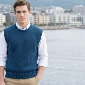 May include: A man wearing a blue knitted vest over a white collared shirt and beige trousers. The vest has a ribbed hem and crew neck. The background shows a waterfront with buildings.