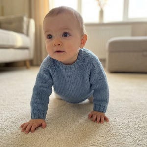 May include: A baby wearing a light blue knitted sweater crawls on a light-colored carpet. The sweater has long sleeves and a crew neck. The baby is looking towards the left side of the frame.