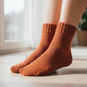 Pu&ograve; includere: Un paio di calzini lavorati a maglia color ruggine. I calzini hanno polsini a coste e un motivo a maglia strutturato. I calzini sono indossati all'interno, con una finestra e una pianta in vaso sullo sfondo. I calzini sembrano essere fatti di un materiale caldo e morbido.