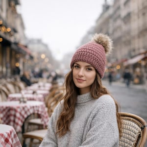 Peut inclure: Une femme porte un bonnet en tricot mauve avec un pompon en fausse fourrure. Elle porte également un pull à col roulé gris. L'arrière-plan montre une scène de rue floue avec des sièges de café en plein air.