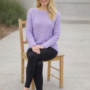 May include: A lilac knitted sweater with a textured design. The sweater features a boat neck and long sleeves. Worn with black leggings. The person is seated on a wooden chair.