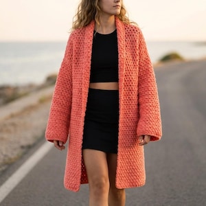 May include: A coral-colored, open-front crochet cardigan. The long-sleeved sweater is knee-length and has a loose, textured weave. The person in the image is wearing a black top and skirt. The background is a road and the ocean.