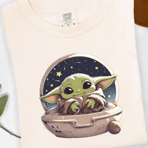 Puede incluir: Camiseta color crema con una imagen de dibujos animados de un personaje verde con aspecto de extraterrestre en una c&aacute;psula espacial sobre un fondo estrellado. La camiseta tiene una etiqueta Comfort Colors y est&aacute; doblada. Un sombrero marr&oacute;n es visible en el fondo.