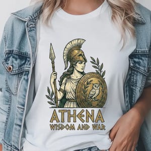 May include: A white t-shirt featuring a graphic illustration of the Greek goddess Athena wearing a helmet, holding a spear, and carrying a shield with an owl. The text below the illustration reads ATHENA WISDOM AND WAR in gold lettering.