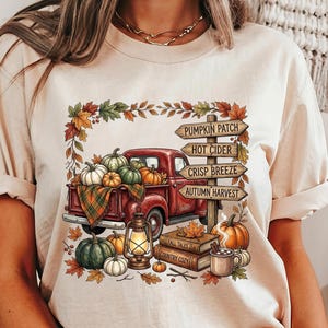 Puede incluir: Una camiseta beige con un gr&aacute;fico de una camioneta roja cargada de calabazas, una linterna y libros. Un poste de madera muestra el texto PUMPKIN PATCH, HOT CIDER, CRISP BREEZE y AUTUMN HARVEST, rodeado de hojas de oto&ntilde;o.