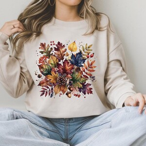 May include: A beige crewneck sweatshirt featuring a central watercolor-style graphic of autumn foliage, including various colorful leaves, pinecones, acorns, and small red berries arranged in a dense, decorative cluster on the front chest area.