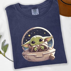 Puede incluir: Camiseta azul marino con una ilustraci&oacute;n de dibujos animados de un personaje alien&iacute;gena verde en una c&aacute;psula contra un fondo estrellado. La camiseta est&aacute; hecha de un material suave y tiene la etiqueta de la marca Comfort Colors.