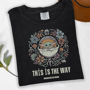 May include: Black t-shirt featuring a floral wreath design with a cartoon image of Grogu from The Mandalorian. The text "THIS IS THE WAY" and "MANDALORIAN GROGU" are printed below the image. The shirt is a Comfort Colors brand.