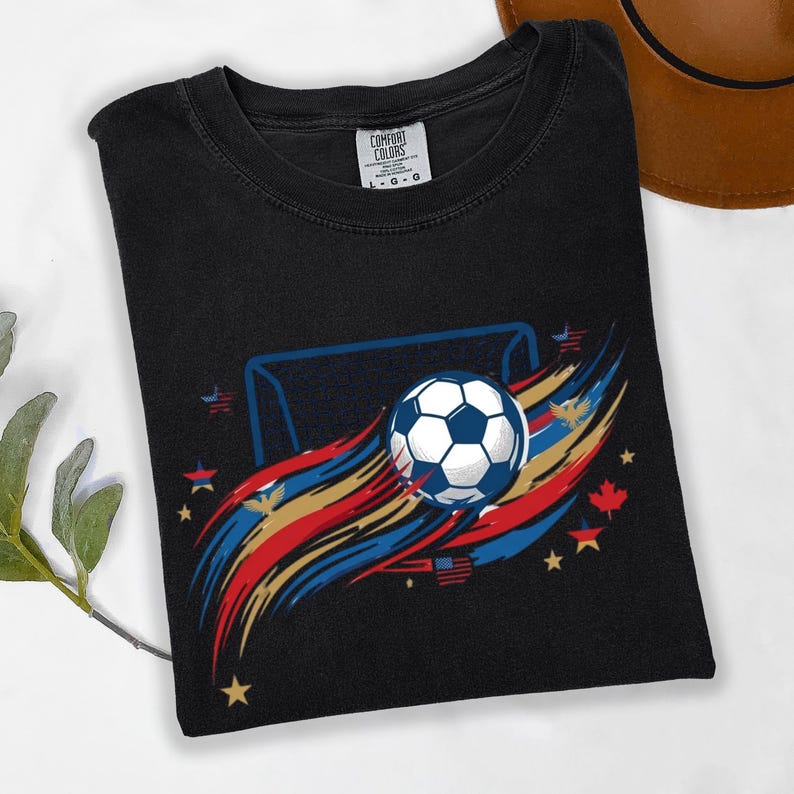 May include: A black t-shirt featuring a graphic of a soccer ball in front of a goal, surrounded by red, gold, and blue brushstroke accents, small stars, eagles, and maple leaf icons. The inner neck label reads Comfort Colors size L.