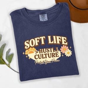 May include: A navy blue t-shirt with the words "SOFT LIFE HUSTLE CULTURE Protect Your Peace" in a retro design. The graphic includes a sun, flowers, and stars. The shirt is folded and has a Comfort Colors tag.