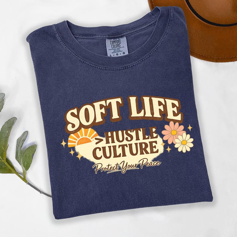 May include: A navy blue t-shirt with the words "SOFT LIFE HUSTLE CULTURE Protect Your Peace" in a retro design. The graphic includes a sun, flowers, and stars. The shirt is folded and has a Comfort Colors tag.
