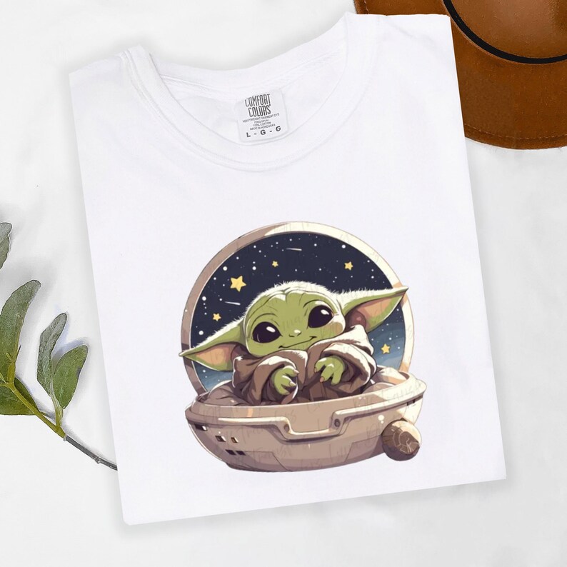 Puede incluir: Camiseta blanca con una ilustraci&oacute;n de dibujos animados de un personaje alien&iacute;gena verde en una c&aacute;psula sobre un fondo estrellado. El personaje tiene grandes ojos negros y orejas puntiagudas. La camiseta tiene cuello redondo y la marca "Comfort Colors" es visible.